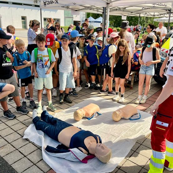 Eine Gruppe von Kindern und Erwachsenen versammelt sich um einen Tisch mit CPR-Manequins, wobei eine Person in roter und gelber Sicherheitsausrüstung in der Nähe steht.