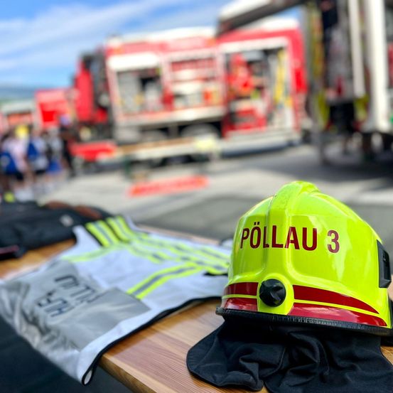 Ein gelber Helm mit der Aufschrift Pollau 3 steht auf einem Holztisch mit einer reflektierenden Weste und Handschuhen. Im Hintergrund sind mehrere Feuerwehrfahrzeuge geparkt.