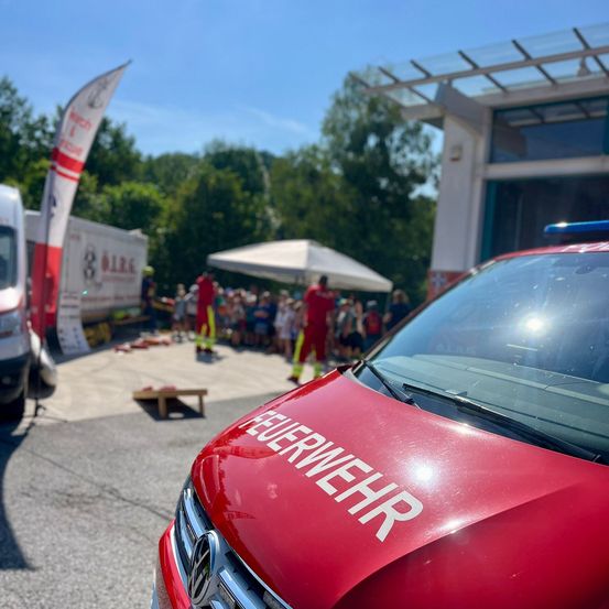 Ein roter Feuerwehrwagen steht vor einem Gebäude, während eine Menschenmenge eine Veranstaltung beobachtet. Das Wort 'Feuerwehr' ist auf der Vorderseite des Wagens geschrieben.