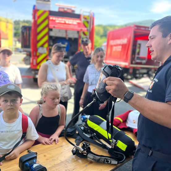 Ein Feuerwehrmann demonstriert Ausrüstung für eine Gruppe von Kindern und Erwachsenen in einer Außenumgebung mit Feuerwehrwagen im Hintergrund.