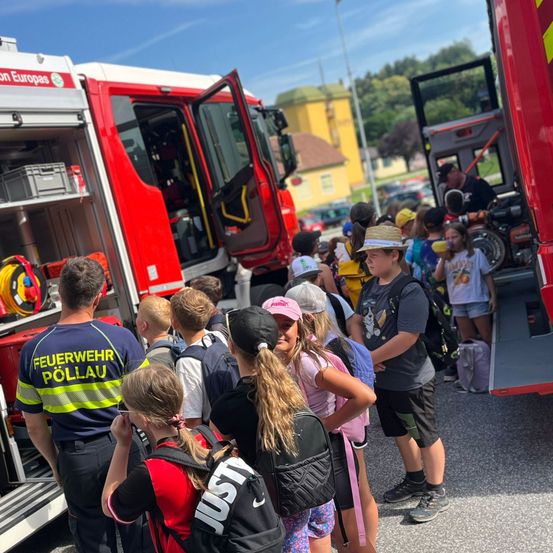 Eine Gruppe von Kindern und Erwachsenen versammelt sich um einen Feuerwehrwagen, wobei ein Erwachsener in Uniform vor dem Fahrzeug steht.