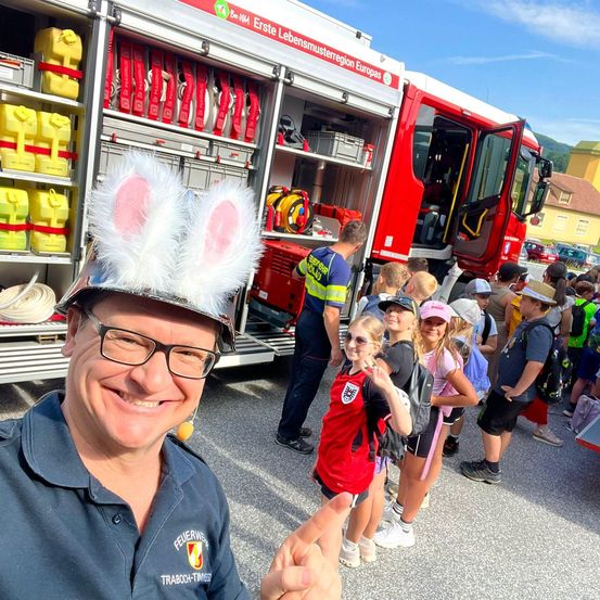 Ein Feuerwehrmann mit Hasenohren posiert mit einer Gruppe von Kindern vor einem Feuerwehrauto. Die Kinder tragen Rucksäcke und einige haben Hüte.