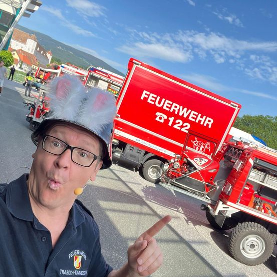 Ein Mann mit einem Feuerwehrhelm mit Hasenohren zeigt auf einen roten Feuerwehrwagen mit der Aufschrift 'Feuerwehr' und '122'. Der Wagen steht auf einer Straße, im Hintergrund sind weitere Fahrzeuge zu sehen.