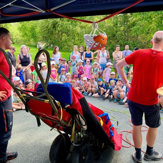 Eine Gruppe von Kindern und Erwachsenen beobachtet, wie ein Mann einen roten Gurt aus einem Trolley unter einem Zelt hebt.