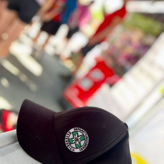Nahaufnahme eines schwarzen Baseball-Caps mit einem Logo, das ein grünes Kreuz und den Text 'TISSO BERGGESETTUNGEN' zeigt. Der Hintergrund ist unscharf, mit Menschen, die in der Ferne gehen.