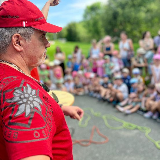 Ein Mann in einem roten Shirt und einer roten Kappe steht vor einer Gruppe von Kindern, möglicherweise in einem Sommercamp. Die Kinder sitzen im Kreis auf dem Rasen.
