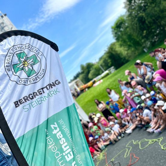 Eine Menschenmenge, einschließlich Kinder, versammelt sich auf einem Rasenfeld, möglicherweise für eine Veranstaltung. Ein Banner mit dem Text 'BERGRETTUNG STEIERMARK' ist im Vordergrund sichtbar.