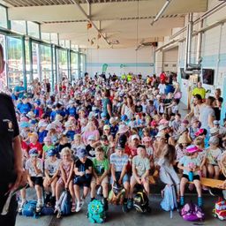 Eine große Gruppe von Kindern und Erwachsenen versammelt sich in einer Sporteinrichtung, viele tragen Hüte und haben Rucksäcke dabei. Der Raum ist gut beleuchtet mit großen Fenstern und hat eine Mischung aus Sitz- und Stehbereichen.