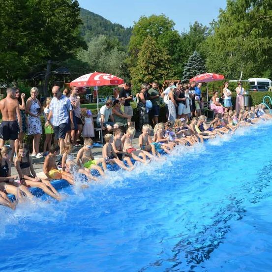 Eine Gruppe von Kindern und Erwachsenen steht in einem Pool, einige Erwachsene stehen daneben und schauen zu. Der Pool ist von Bäumen umgeben und in der Nähe befinden sich Regenschirme.