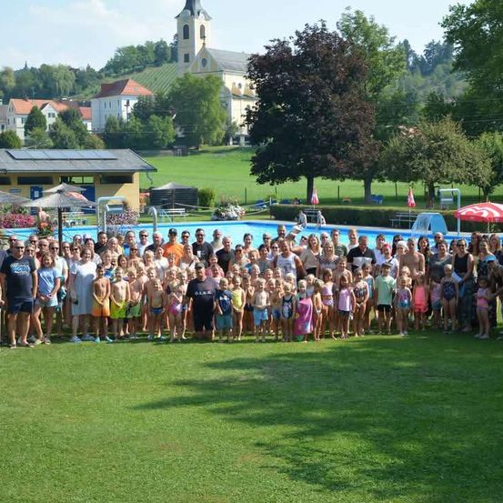 Eine große Gruppe von Menschen, einschließlich Erwachsener und Kinder, versammelt sich auf einer Rasenfläche in der Nähe eines Schwimmbeckens, mit einer Kirche und Häusern im Hintergrund.