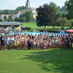 Eine große Gruppe von Menschen, einschließlich Erwachsener und Kinder, versammelt sich auf einer Rasenfläche in der Nähe eines Schwimmbeckens, mit einer Kirche und Häusern im Hintergrund.