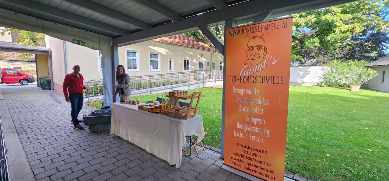 Eine Frau steht neben einem Tisch mit verschiedenen Honigprodukten, vor einem Haus mit Garten. Ein orangefarbener Banner mit einem Männergesicht und Text befindet sich in der Nähe.