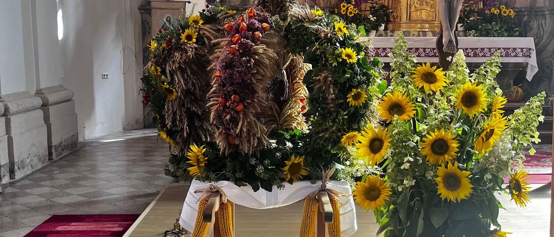 Ein Kircheninneres mit einer Blumenarrangement vor dem Altar, Sonnenblumen und einem Kruzifix. Statuen und Gemälde schmücken die Wände.