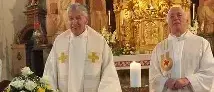 Zwei Priester stehen lächelnd am Altar, mit einer Kerze und Blumen. Sie tragen weiße Gewänder und stehen in einer Kirche mit einem kunstvollen Hintergrund.