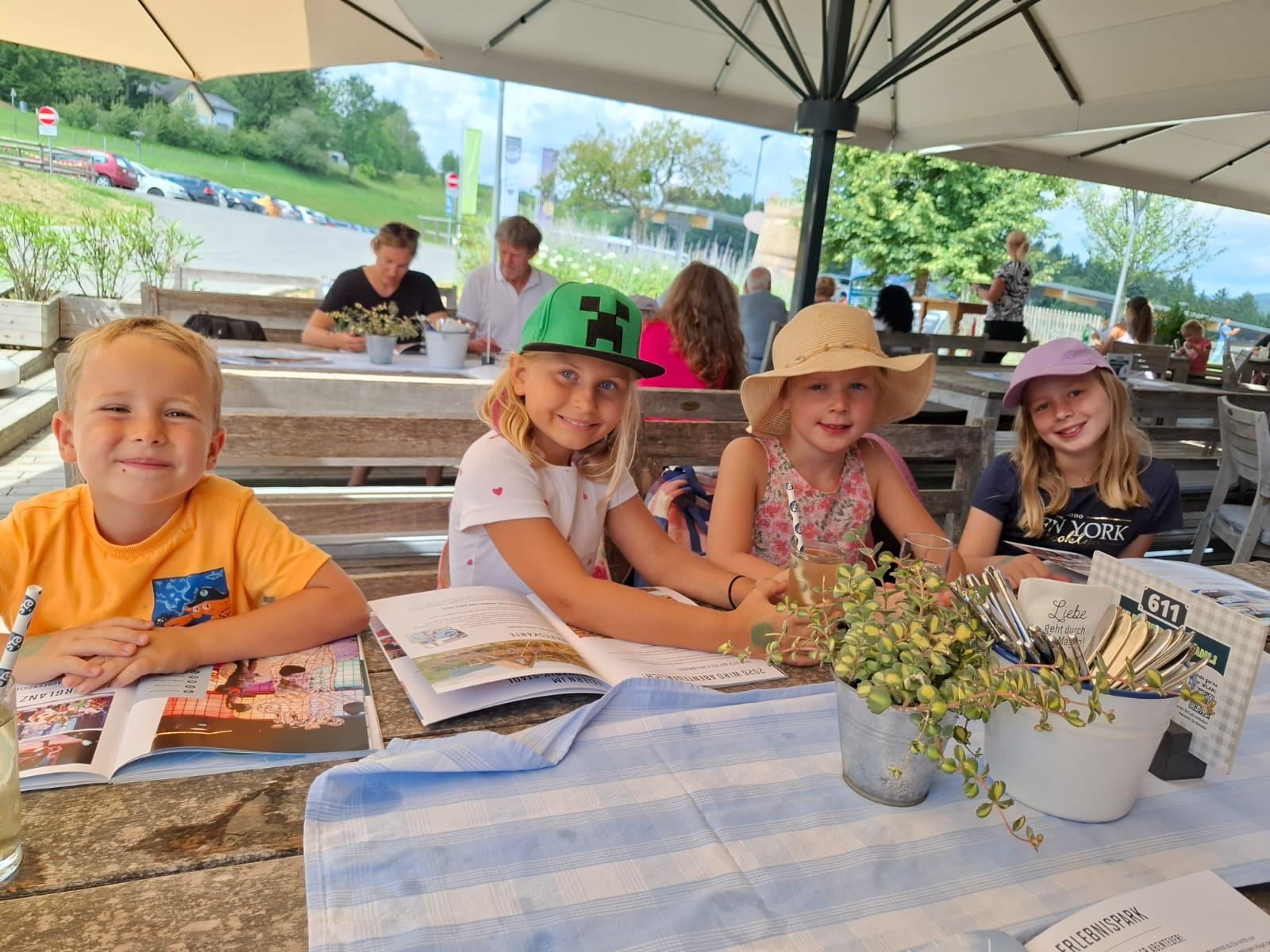 Vier Kinder sitzen unter einem Schirm an einem Tisch und lächeln. Ein Mädchen mit grüner Mütze und ein anderes mit rosa Mütze lesen Bücher. Ein Topf mit Pflanzen und Besteck sind auf dem Tisch.