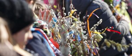 A group of children and adults hold decorative branches with colorful ribbons. The children are wearing winter clothes and hats.