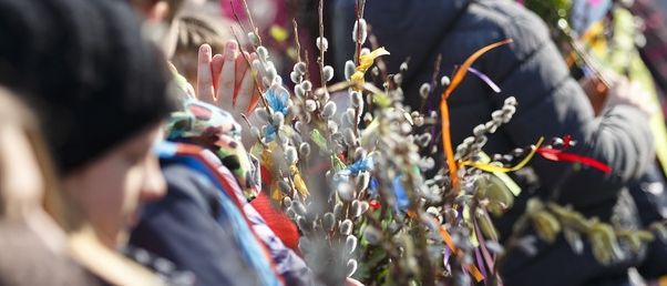 A group of children and adults hold decorative branches with colorful ribbons. The children are wearing winter clothes and hats.