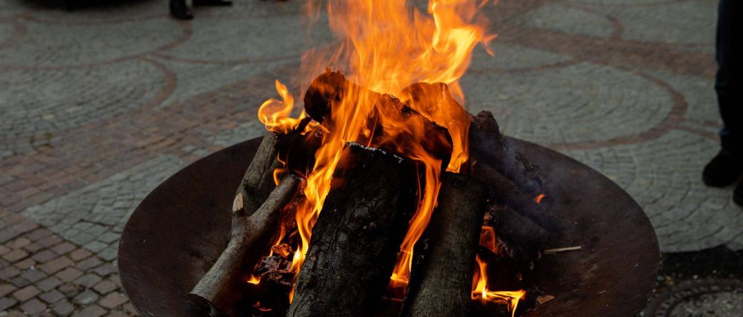 Ein Feuerkorb mit brennendem Holz und Flammen in einer Metallschale auf einem Ziegelboden, mit zwei Personen im Hintergrund.