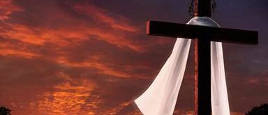 Ein weißes Tuch über einem Kreuz gegen einen leuchtend orangefarbenen Sonnenuntergang mit Baumsilhouetten im Hintergrund.