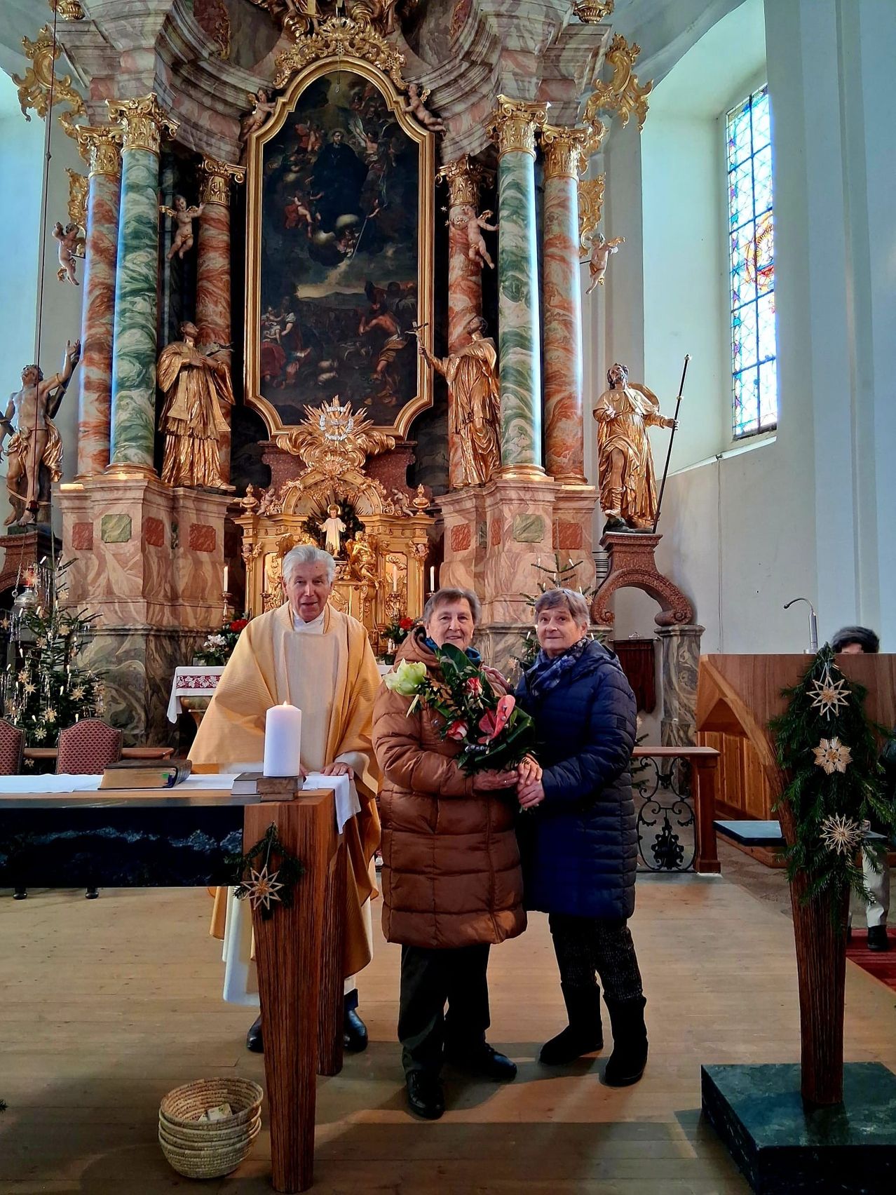 Ein Priester und zwei Frauen stehen in einer Kirche mit einer Kerze und Blumen. Dahinter befindet sich ein geschmückter Altar und Statuen.