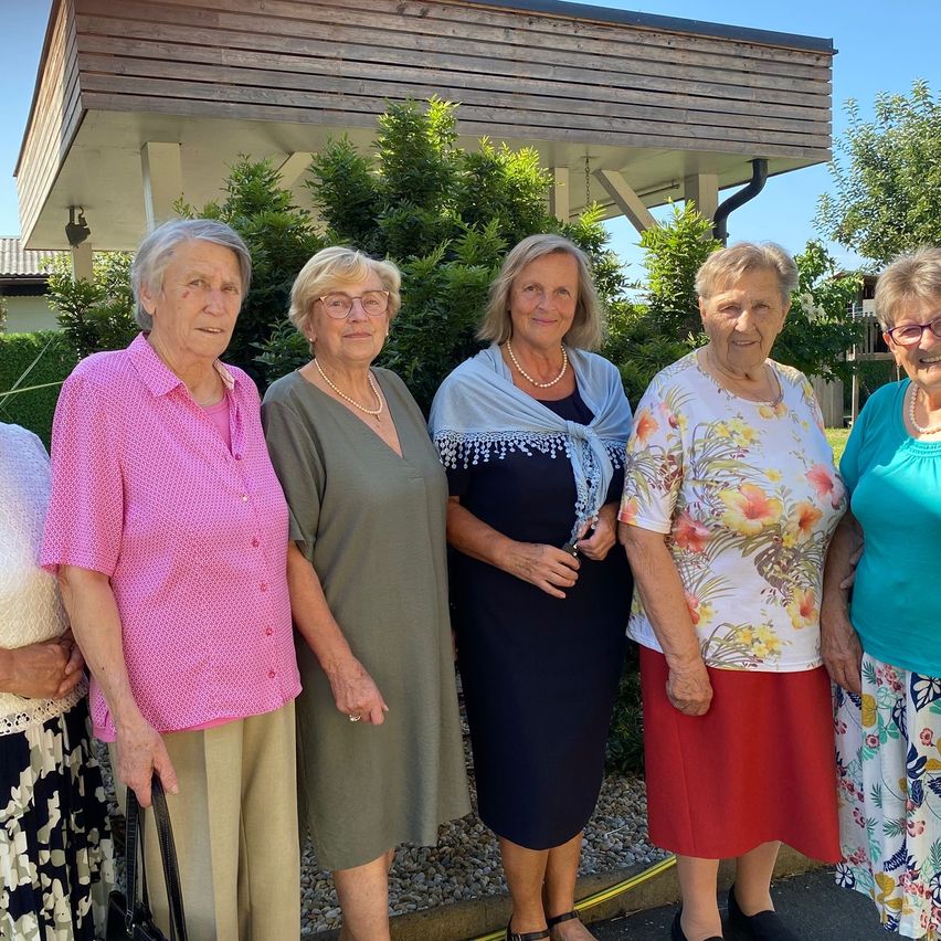 Eine Gruppe älterer Frauen steht draußen und lächelt für ein Foto vor einem Haus mit Holzfassade.