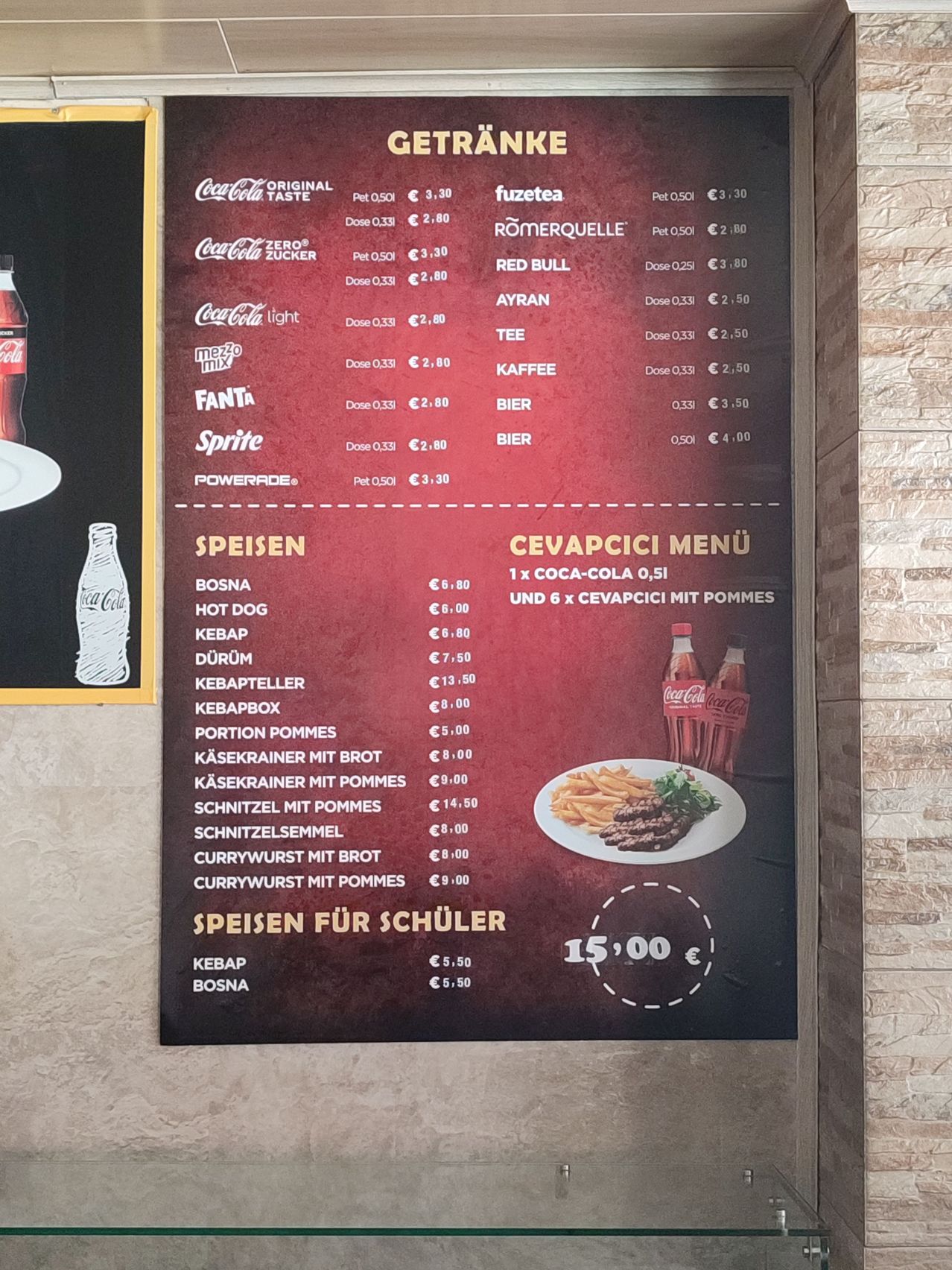 Ein Menü mit Getränken, Snacks und Mahlzeiten. Coca-Cola, Fanta, Sprite und Powerade sind in verschiedenen Größen erhältlich. Zu den Getränken gehören Fuzetea, Romerquelle, Red Bull, Ayran, Tee, Kaffee, Bier. Zu den Speisen gehören Bosna, Hot Dog, Kebab, Dürum, Kebapteller, Kebapbox, Portion Pommes, Käsekrainer mit Brot, Käsekrainer mit Pommes, Schnitzel mit Pommes, Schnitzelsemmel, Currywurst mit Brot, Currywurst mit Pommes. Es gibt auch ein spezielles Menü für Schüler mit Kebab und Bosna.