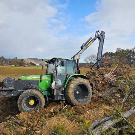 Ein grüner Traktor mit angehängtem Holzharvester arbeitet auf einem Feld mit Erde und umgestürzten Ästen.