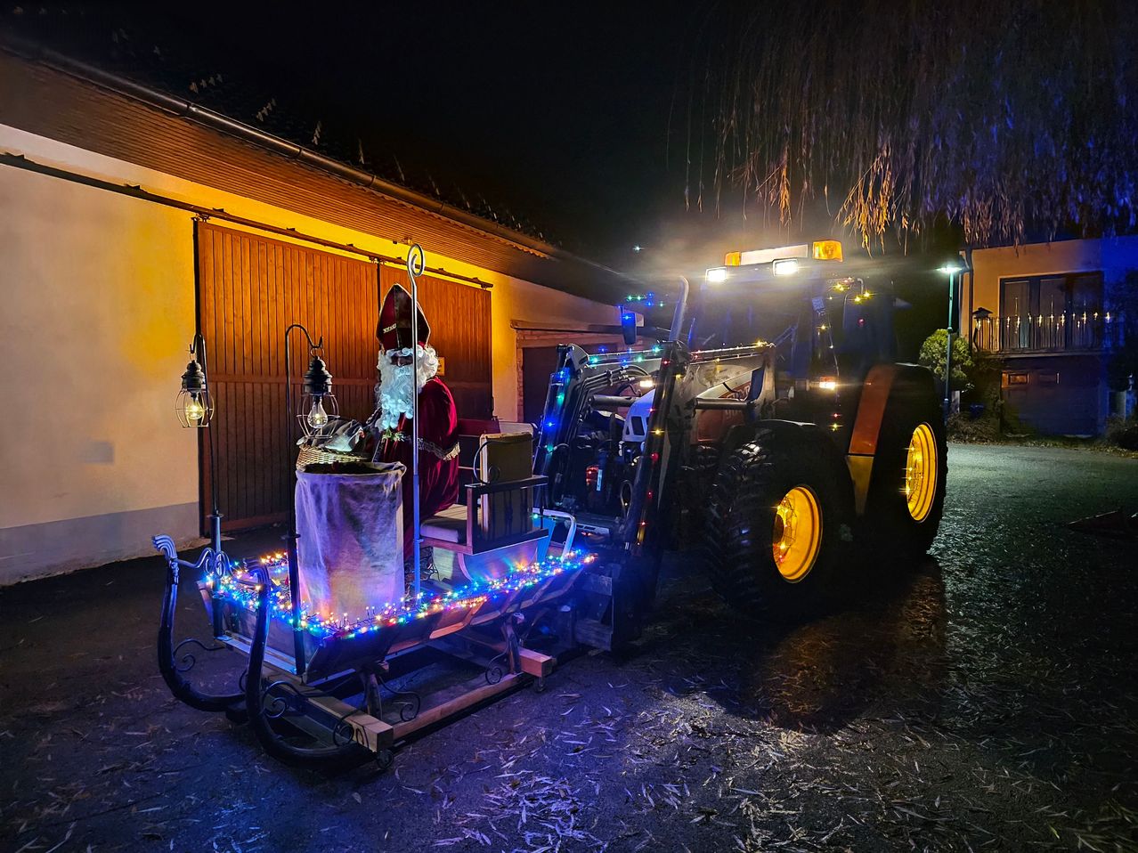 Der Weihnachtsmann sitzt auf einem dekorierten Traktor, der nachts vor einem Gebäude geparkt ist. Der Traktor hat Weihnachtslichter und einen Sack vor dem Weihnachtsmann.