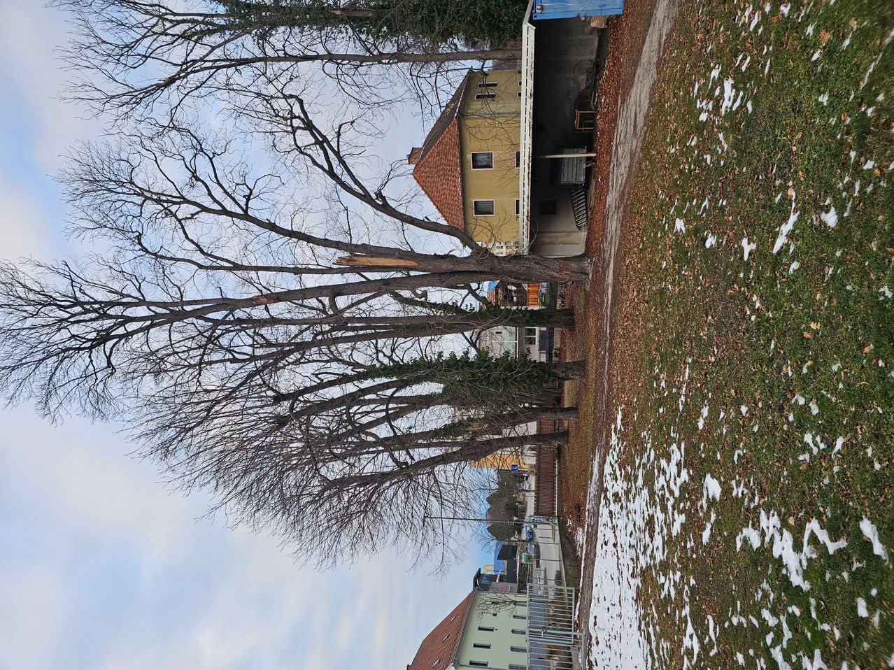 Luftaufnahme eines Hauses mit rotem Dach, umgeben von Bäumen und schneebedecktem Boden. Das Haus hat mehrere Fenster und einen kleinen Balkon. Ein blauer Fass steht in der Nähe des Eingangs.