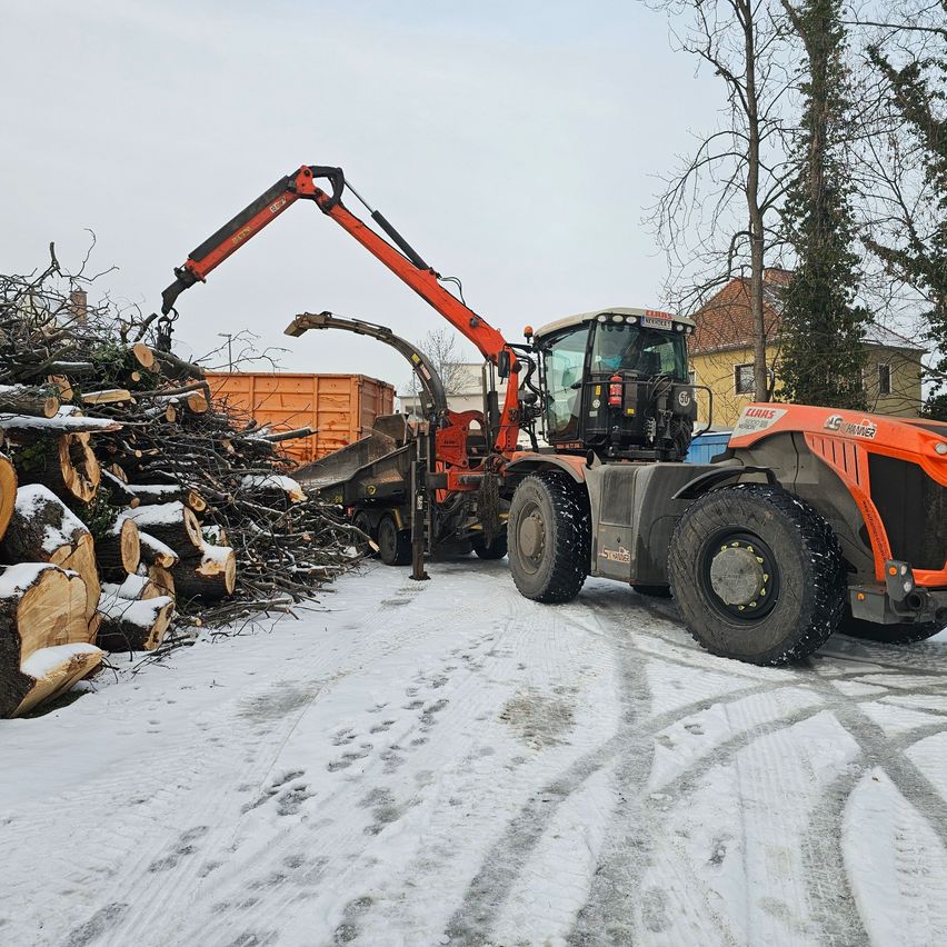 An orange harvester with a crane arm is positioned on a snowy road, loading wood into a truck. Trees with no leaves and houses are visible in the background.
