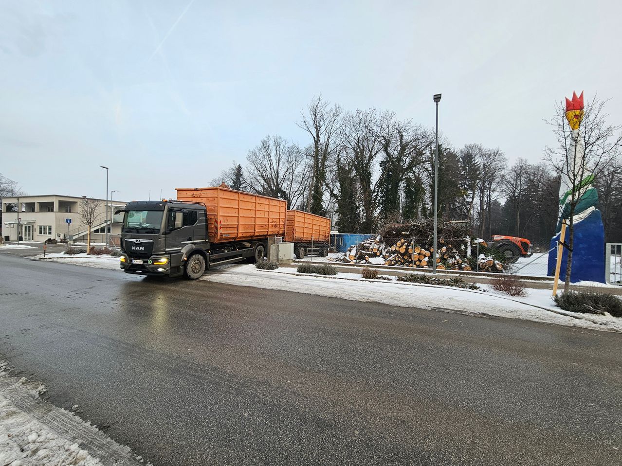 A large black truck with orange trailers is parked on a snowy road. Behind it, there are piles of wood and a fence. The sky is overcast.