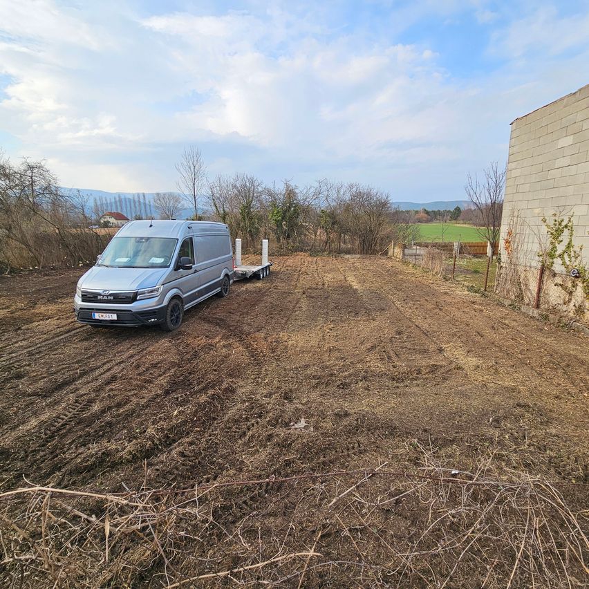 Ein silberner Lieferwagen ist auf einer unbefestigten Straße geparkt, mit einem Anhänger, in der Nähe eines Feldes und eines Gebäudes.