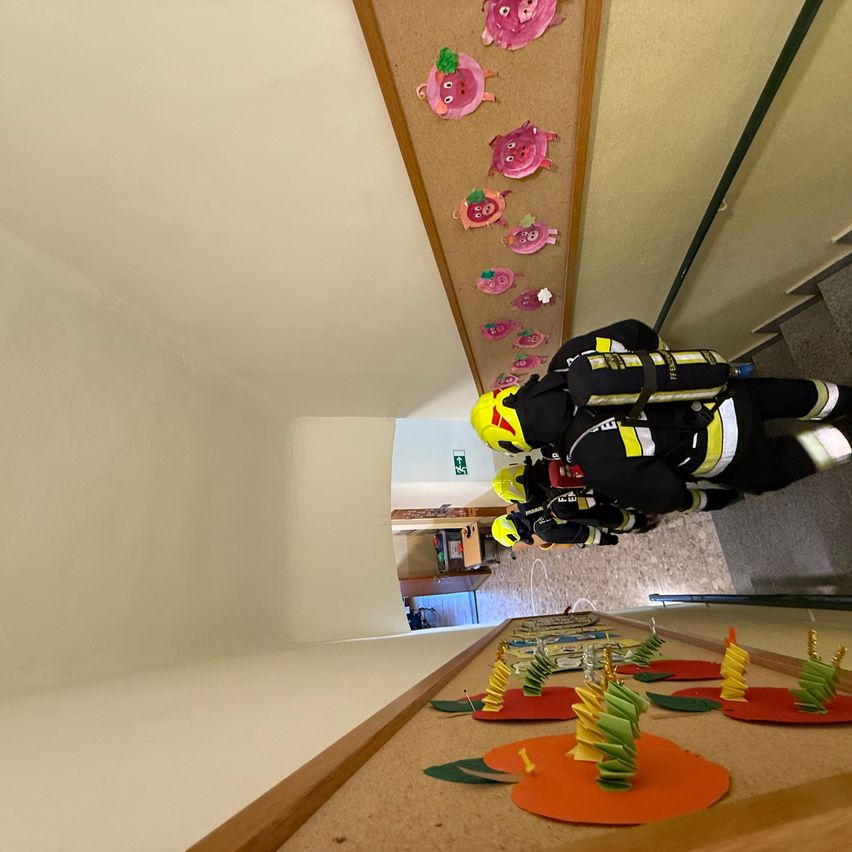 Feuerwehrleute in voller Ausrüstung gehen eine Treppe hinunter, wahrscheinlich zur Übung einer Evakuierung. Die Wand hat bunte Schweineaufkleber und der Boden hat spiralförmige Apfeldekorationen.