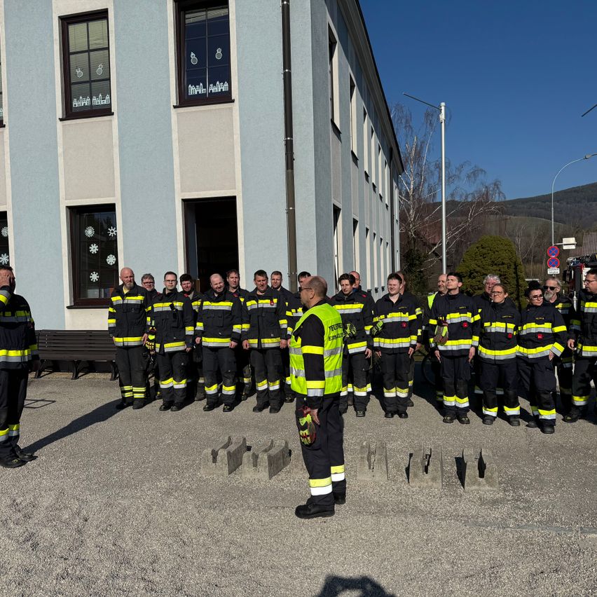 Eine Gruppe Feuerwehrleute in Uniform steht vor einem Gebäude, ein Mann spricht zu ihnen.