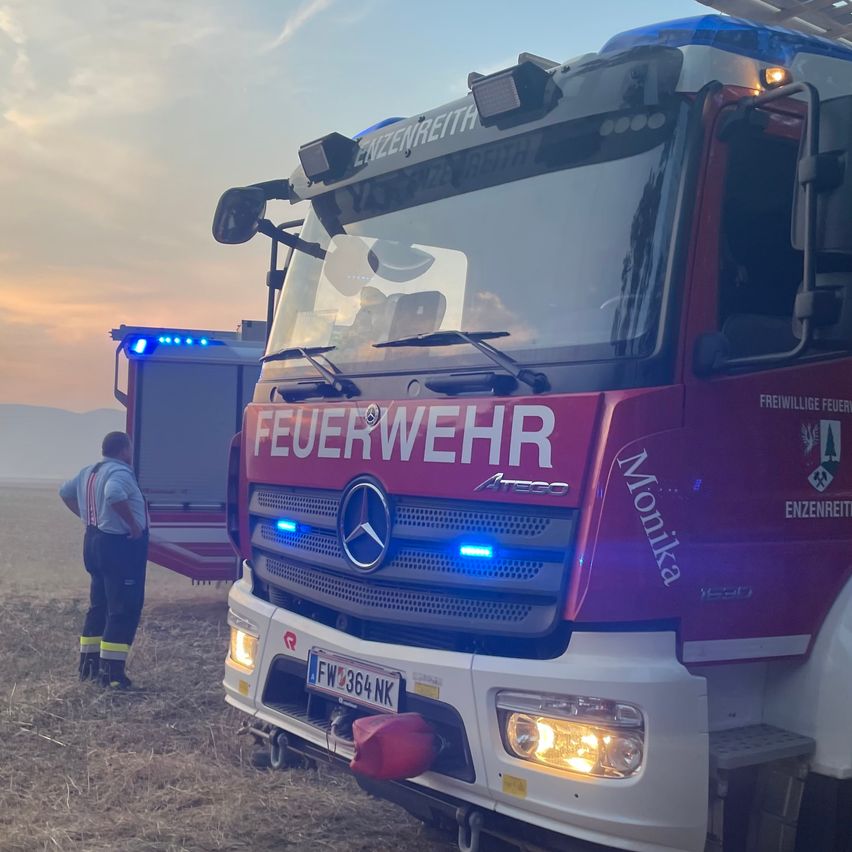 Ein Feuerwehrauto mit dem Wort 'Feuerwehr' auf der Front ist auf einem Feld geparkt. Eine Person in einer reflektierenden Weste steht daneben.
