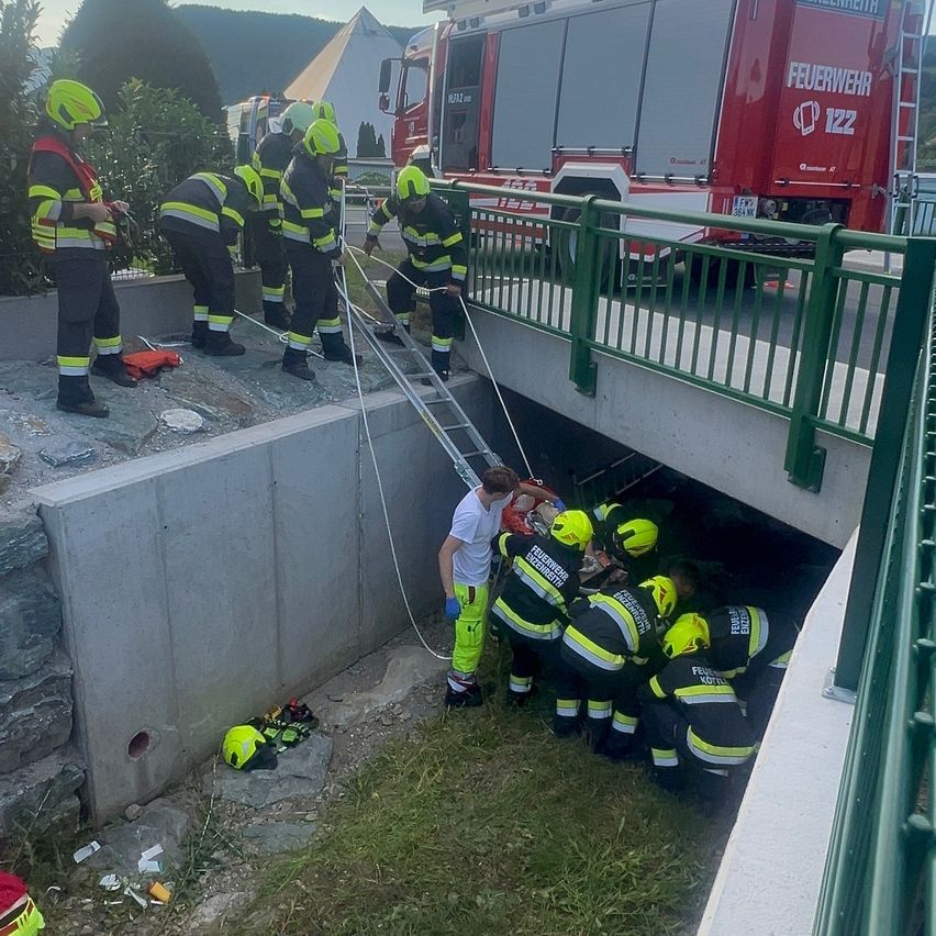 Feuerwehrleute in Schutzkleidung retten jemanden unter einer Brücke. Ein roter Feuerwehrwagen steht in der Nähe.