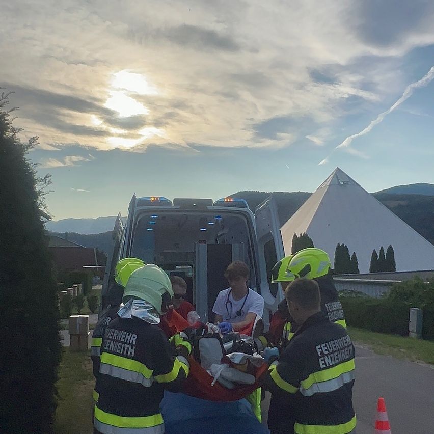 Rettungskräfte entladen einen Patienten aus einem Krankenwagen. Der Patient liegt auf einer Trage, und die Helfer tragen Schutzausrüstung. Die Szene spielt im Freien mit einem pyramidenförmigen Gebäude im Hintergrund.