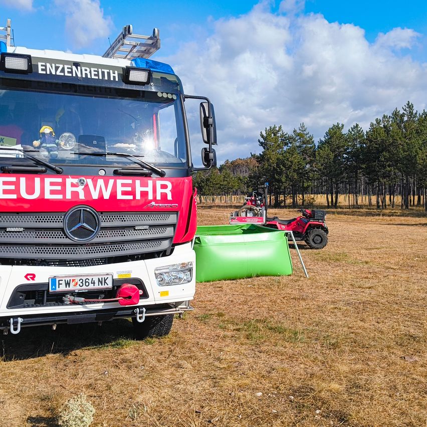 Ein roter Feuerwehrwagen steht auf einem Feld neben einem grünen aufblasbaren Pool. Ein roter ATV ist hinter dem Wagen geparkt. Bäume sind im Hintergrund.