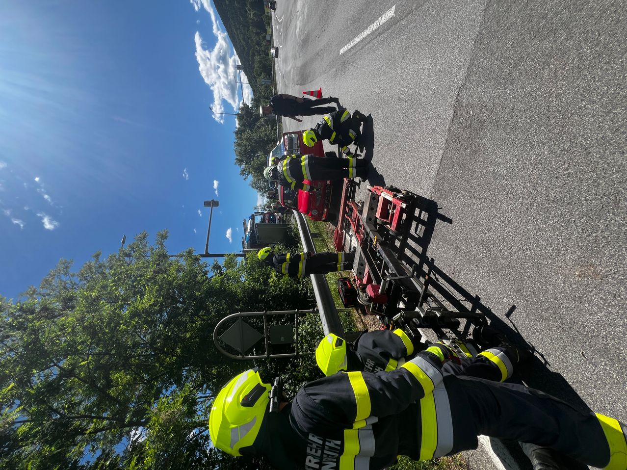 Firefighters in yellow helmets are on the roadside with equipment. They seem to be working on a vehicle, possibly a car. There are orange traffic cones on the road. Trees and bushes are on the side of the road.