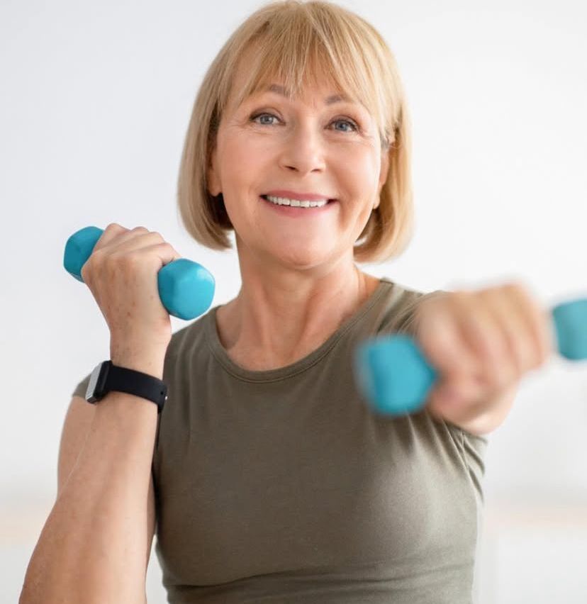 Eine ältere Frau mit kurzen Haaren lächelt, während sie mit blauen Hanteln trainiert. Sie trägt ein braunes Tanktop und eine schwarze Armbanduhr.
