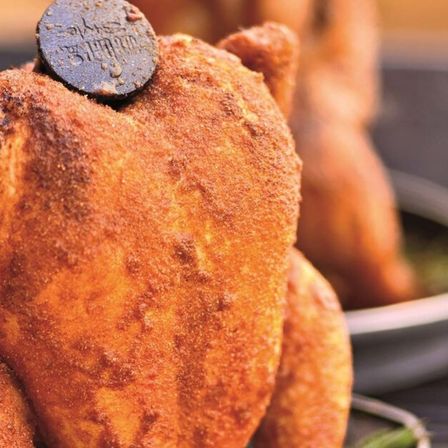 Ein gebratenes Huhn mit knuspriger Haut steht auf einem Tisch, mit einem runden Gegenstand darauf. Das Huhn ist goldbraun.