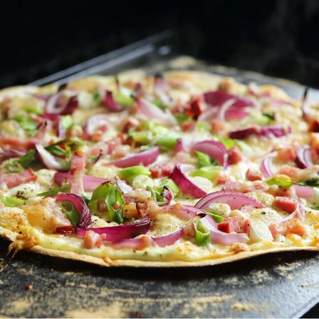 Eine Pizza mit geschmolzenem Käse, roten Zwiebeln, Paprika und Schinkenscheiben, auf einem Backblech zum Backen bereit.