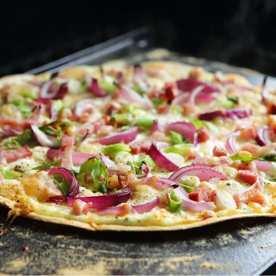 Eine Pizza mit geschmolzenem Käse, roten Zwiebeln, Paprika und Schinkenscheiben, auf einem Backblech zum Backen bereit.