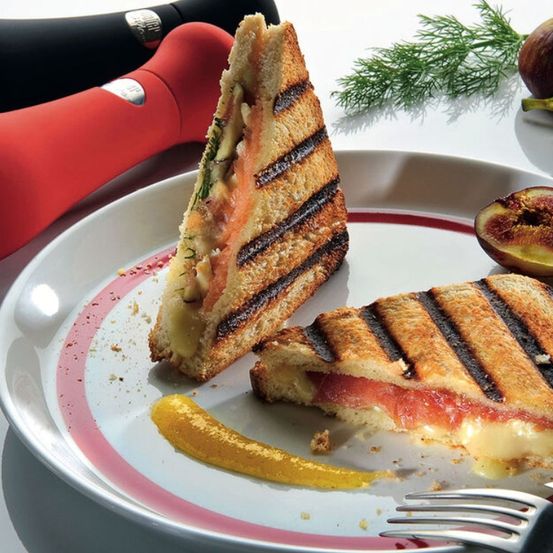 Zwei gegrillte Käsesandwiches auf einem Teller, mit einer Gabel und einer Gurke und Feigen auf der Seite.