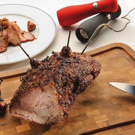 Ein großes gebratenes Schweinebein auf einem Holzschneidebrett mit einem Messer, einer roten und schwarzen Pfeffermühle und einem Teller mit Fleisch.