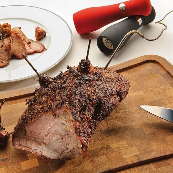 Ein großes gebratenes Schweinebein auf einem Holzschneidebrett mit einem Messer, einer roten und schwarzen Pfeffermühle und einem Teller mit Fleisch.