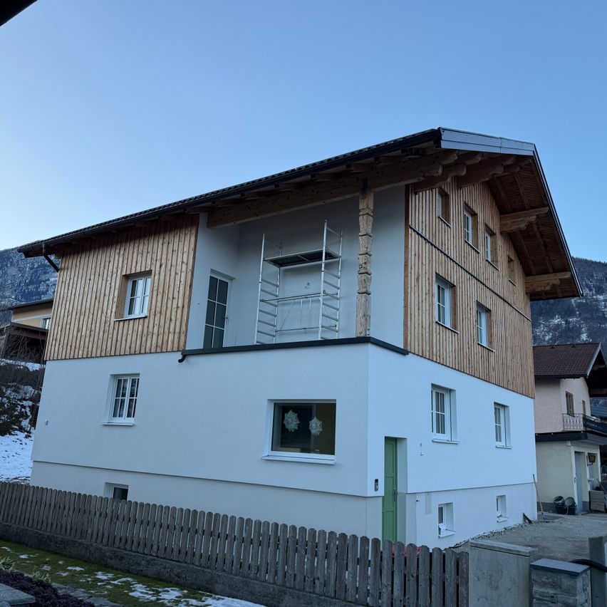 Ein dreistöckiges Haus mit Holzakzenten und einem grauen Dach. Es hat einen Holzzaun und Fenster, mit einer Leiter und einem Balkon. Im Hintergrund befindet sich ein verschneiter Berg.