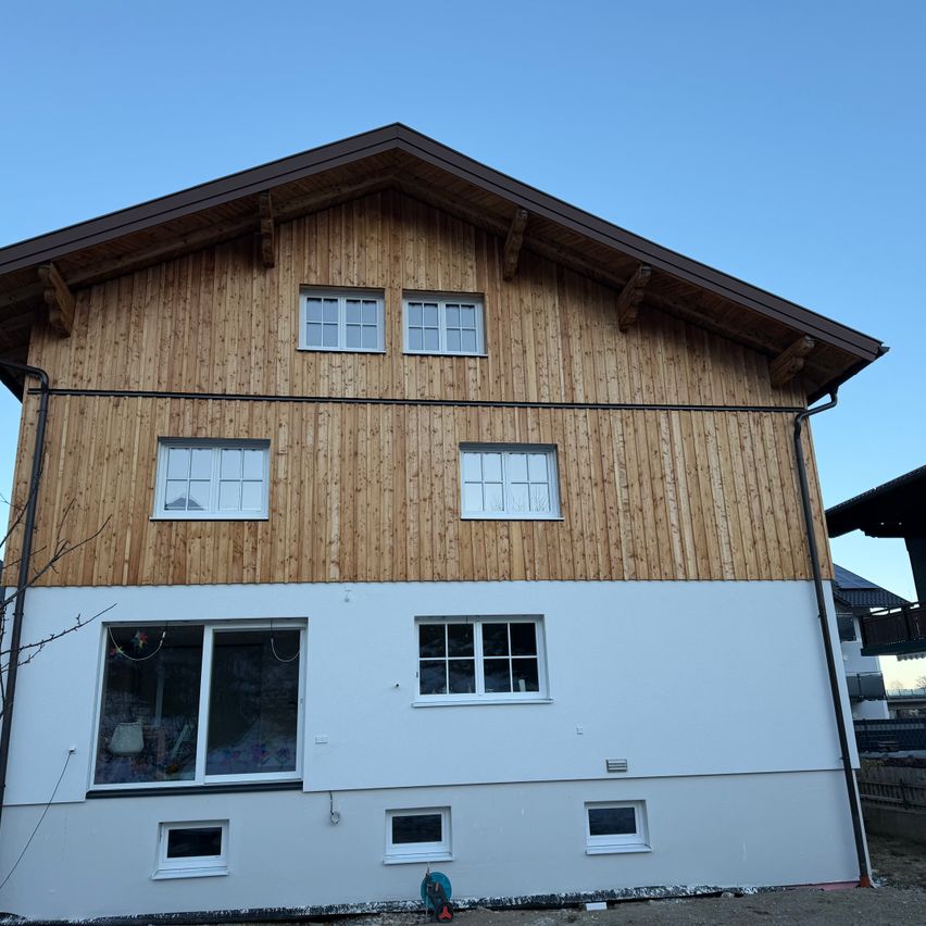 Ein modernes Haus mit einer Holzfassade und weißen Wänden, mit mehreren Fenstern und einem Schornstein auf dem Dach.