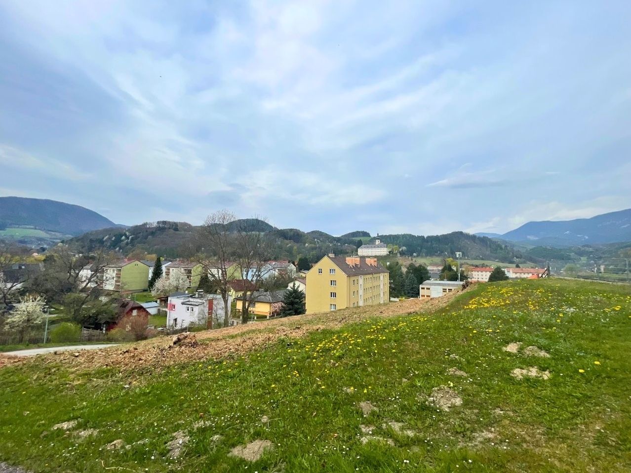 A hillside view of a village with several buildings, some with yellow walls, and a mountain range in the background under a partly cloudy sky.