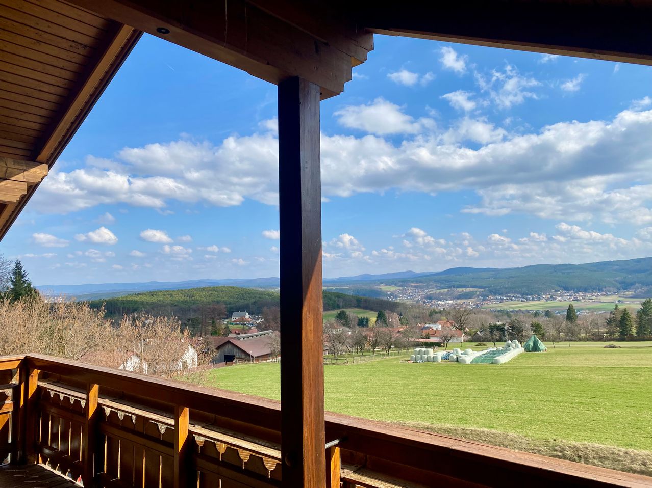 Der Balkonblick aus einer Holzstruktur zeigt einen sonnigen Tag mit blauem Himmel und weißen Wolken. Im Vordergrund befinden sich ein grünes Feld, entfernte Gebäude und Bäume. Am Horizont sind Berge sichtbar.